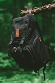 A black backpack hangs from a tree branch in a lush, green forest setting. The backpack features a leather patch and multiple compartments, with a focus on its sturdy design.