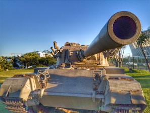 A large military tank is prominently positioned in an outdoor setting, with its massive barrel extending toward the viewer. The tank is surrounded by grass and trees, and a modern building is visible in the background under a clear blue sky.