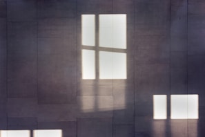 Light filtering through window panes casts shadows on a tiled wall, creating a geometric and minimalist visual effect.