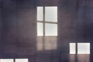 Light filtering through window panes casts shadows on a tiled wall, creating a geometric and minimalist visual effect.