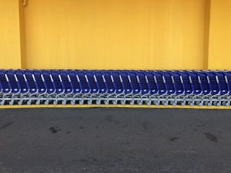A row of shopping carts arranged neatly in front of a vibrant yellow wall. The carts are blue with metallic frames, and they are aligned side by side on a dark gray pavement.