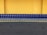 A row of shopping carts arranged neatly in front of a vibrant yellow wall. The carts are blue with metallic frames, and they are aligned side by side on a dark gray pavement.