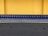 A row of shopping carts arranged neatly in front of a vibrant yellow wall. The carts are blue with metallic frames, and they are aligned side by side on a dark gray pavement.