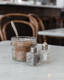 A glass jar filled with brown sugar, featuring a spoon inside, is placed on a white marble table. Next to it, there are two vintage-style glass shakers, likely for salt and pepper, with metal lids. The table is in a setting with wooden chairs in the background, creating a cozy and classic ambiance.