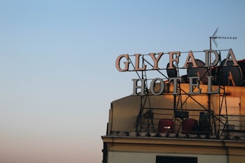 Large metal letters spelling 'GLYFADA HOTEL' are mounted on top of a building. The structure is outlined against a gradient sky that fades from blue to pink. Several satellite dishes and an antenna are visible on the roof.