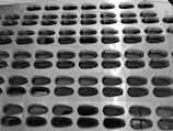 A modern architectural facade with a grid of oval-shaped reflective windows arranged in a repetitive pattern. The surface appears metallic and slightly glossy, with a sleek, futuristic design.