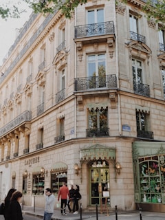 A classic Parisian building with ornate balconies and decorative stone facades houses a Ladurée store. The entrance is adorned with elegant lighting and gold lettering. People are walking on the sidewalk, adding a sense of a lively urban atmosphere. The windows display intricate patterns, enhancing the building's historic charm.