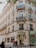 A classic Parisian building with ornate balconies and decorative stone facades houses a Ladurée store. The entrance is adorned with elegant lighting and gold lettering. People are walking on the sidewalk, adding a sense of a lively urban atmosphere. The windows display intricate patterns, enhancing the building's historic charm.