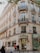 A classic Parisian building with ornate balconies and decorative stone facades houses a Ladurée store. The entrance is adorned with elegant lighting and gold lettering. People are walking on the sidewalk, adding a sense of a lively urban atmosphere. The windows display intricate patterns, enhancing the building's historic charm.