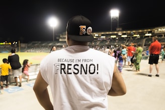 A person is wearing a sleeveless white shirt with the text 'Because I'm from Fresno!' standing in a stadium, likely a baseball game. Other people, including a few children, are nearby, looking towards the field. The stadium lights are bright, and the seating and field are visible in the background.