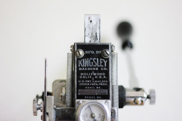 A close-up view of a vintage machine manufactured by Kingsley Machine Co. The machine features metal components, with visible bolts and screws, and includes a label displaying the company's name along with patent details. The background is slightly blurred, emphasizing the focus on the machine.