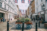 A bustling city street is lined with historic buildings adorned with Union Jack flags. People walk casually on the sidewalk, and a wooden cart filled with colorful plants and flowers placed in the center adds a touch of greenery. The architecture is a blend of white and brown facades, and there's a well-known bank located on the right side. The atmosphere feels vibrant and lively.
