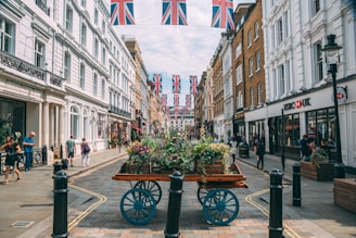 A bustling city street is lined with historic buildings adorned with Union Jack flags. People walk casually on the sidewalk, and a wooden cart filled with colorful plants and flowers placed in the center adds a touch of greenery. The architecture is a blend of white and brown facades, and there's a well-known bank located on the right side. The atmosphere feels vibrant and lively.
