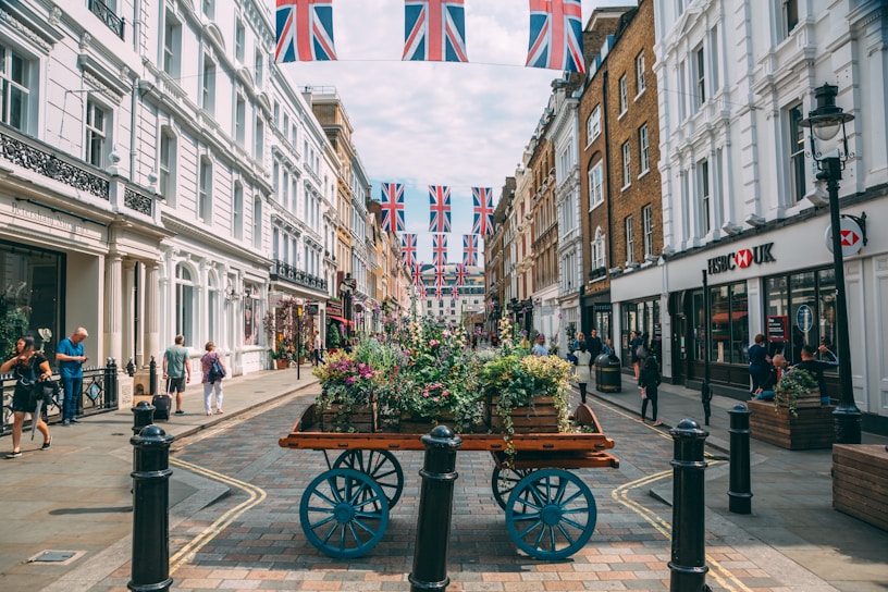 A bustling city street is lined with historic buildings adorned with Union Jack flags. People walk casually on the sidewalk, and a wooden cart filled with colorful plants and flowers placed in the center adds a touch of greenery. The architecture is a blend of white and brown facades, and there's a well-known bank located on the right side. The atmosphere feels vibrant and lively.