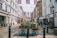 A bustling city street is lined with historic buildings adorned with Union Jack flags. People walk casually on the sidewalk, and a wooden cart filled with colorful plants and flowers placed in the center adds a touch of greenery. The architecture is a blend of white and brown facades, and there's a well-known bank located on the right side. The atmosphere feels vibrant and lively.