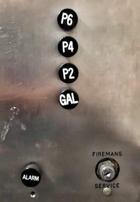 A metallic panel with elevator buttons labeled P6, P4, P2, and GAL in a vertical alignment. Below the buttons, there is a black button labeled ALARM and a keyhole labeled as FIREMANS SERVICE.
