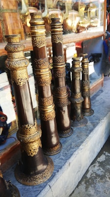 Several intricately designed musical instruments with ornate golden and bronze detailing are arranged on a stone shelf. Behind the instruments, there are reflections and glimpses of other metallic items, possibly more instruments or decorative objects.
