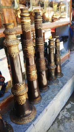 Several intricately designed musical instruments with ornate golden and bronze detailing are arranged on a stone shelf. Behind the instruments, there are reflections and glimpses of other metallic items, possibly more instruments or decorative objects.