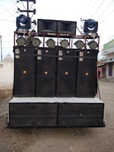 A large sound system mounted on a transportable frame with multiple speakers and lights, located outdoors on a dirt path. The speakers are stacked in columns and rows, with spotlights and stage lights on top.