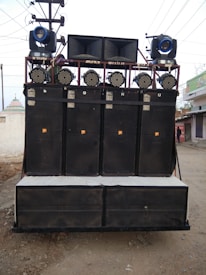 A large sound system mounted on a transportable frame with multiple speakers and lights, located outdoors on a dirt path. The speakers are stacked in columns and rows, with spotlights and stage lights on top.