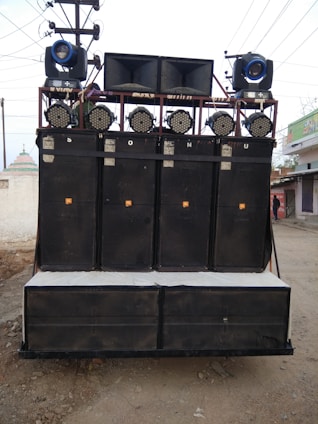 A large sound system mounted on a transportable frame with multiple speakers and lights, located outdoors on a dirt path. The speakers are stacked in columns and rows, with spotlights and stage lights on top.