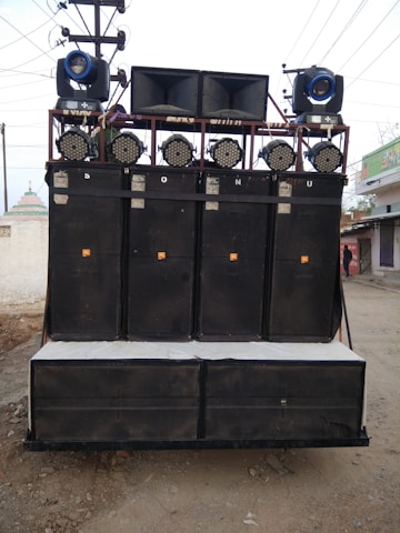 A large sound system mounted on a transportable frame with multiple speakers and lights, located outdoors on a dirt path. The speakers are stacked in columns and rows, with spotlights and stage lights on top.