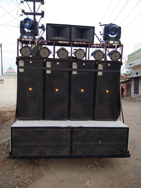 A large sound system mounted on a transportable frame with multiple speakers and lights, located outdoors on a dirt path. The speakers are stacked in columns and rows, with spotlights and stage lights on top.