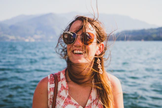 A smiling woman wearing gemlani earrings by the ocean, with soft waves in the background.