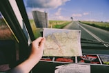 A hand is holding a map inside a car while traveling on a deserted road. The map displays various roads and destinations, suggesting a journey through a rural or remote area. The view through the windshield shows an open road stretching into the horizon, flanked by grassy fields under a partly cloudy sky. Several stickers are visible on the dashboard.
