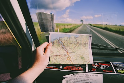A hand is holding a map inside a car while traveling on a deserted road. The map displays various roads and destinations, suggesting a journey through a rural or remote area. The view through the windshield shows an open road stretching into the horizon, flanked by grassy fields under a partly cloudy sky. Several stickers are visible on the dashboard.