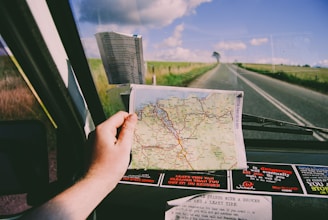 A hand is holding a map inside a car while traveling on a deserted road. The map displays various roads and destinations, suggesting a journey through a rural or remote area. The view through the windshield shows an open road stretching into the horizon, flanked by grassy fields under a partly cloudy sky. Several stickers are visible on the dashboard.