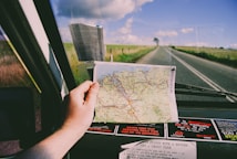 A hand is holding a map inside a car while traveling on a deserted road. The map displays various roads and destinations, suggesting a journey through a rural or remote area. The view through the windshield shows an open road stretching into the horizon, flanked by grassy fields under a partly cloudy sky. Several stickers are visible on the dashboard.