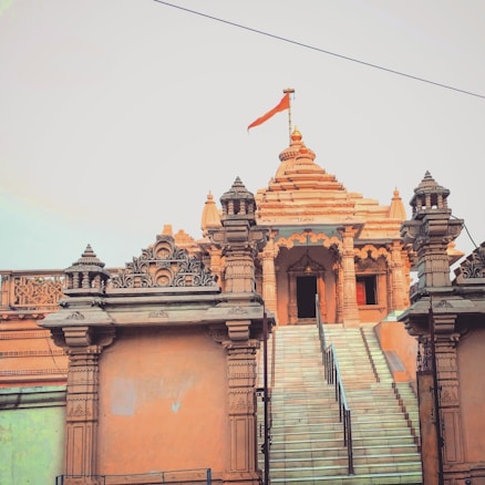 An ornate structure with intricate carvings and a steep staircase leading to the entrance. A striking flag flutters atop the central spire. The architectural style suggests influences from Indian temple design, with detailed patterns and stonework.