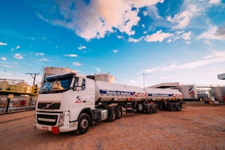 A large white tanker truck with multiple trailers is parked in an industrial area. The truck displays the logo of a company named ALE and carries a label indicating quality control. The surrounding area includes large cylindrical storage tanks and various industrial structures. The sky is bright with patches of clouds, suggesting clear weather.