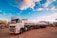 A large white tanker truck with multiple trailers is parked in an industrial area. The truck displays the logo of a company named ALE and carries a label indicating quality control. The surrounding area includes large cylindrical storage tanks and various industrial structures. The sky is bright with patches of clouds, suggesting clear weather.