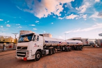 A large white tanker truck with multiple trailers is parked in an industrial area. The truck displays the logo of a company named ALE and carries a label indicating quality control. The surrounding area includes large cylindrical storage tanks and various industrial structures. The sky is bright with patches of clouds, suggesting clear weather.