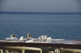 A table set for dining by the seaside, featuring glassware, cutlery, and napkins on placemats. The background shows a calm, expansive body of water under a clear sky.