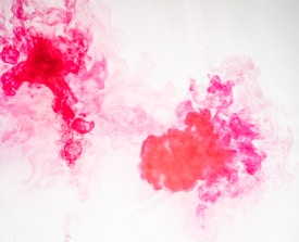 Vibrant red and pink ink swirls through clear water, creating intricate patterns and textures. The wisps and clouds of color intermingle, forming dynamic shapes and soft gradients against a light background.