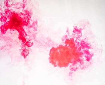 Vibrant red and pink ink swirls through clear water, creating intricate patterns and textures. The wisps and clouds of color intermingle, forming dynamic shapes and soft gradients against a light background.