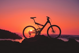 A silhouette of a bicycle is positioned against a vivid, colorful sunset background. The horizon features an orange and pink gradient sky, with the sun setting near the ocean. The bicycle is prominently outlined, adding contrast against the bright backdrop.
