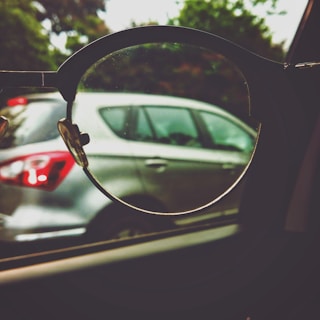 A pair of eyeglasses held up to a car window, focusing on the car outside. The lens frames the vehicle, highlighting the contrast between the lens-focused sharpness and the blurred surroundings.