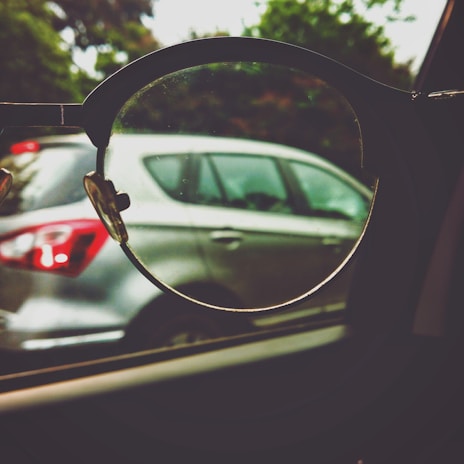 A pair of eyeglasses held up to a car window, focusing on the car outside. The lens frames the vehicle, highlighting the contrast between the lens-focused sharpness and the blurred surroundings.