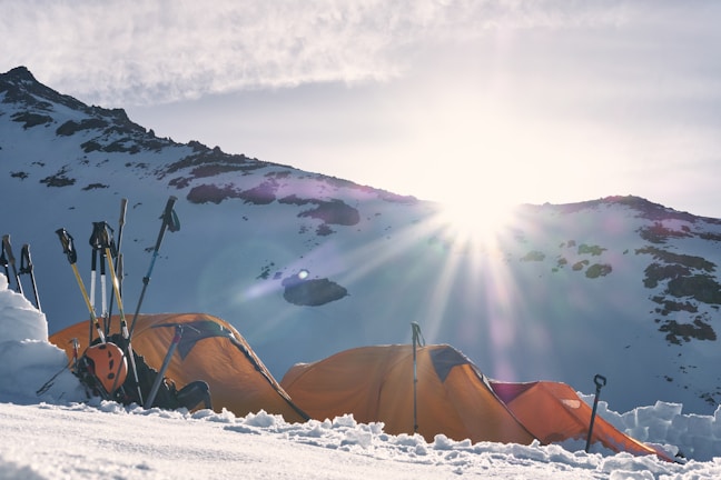 Bright sunlight illuminates snow-covered mountains with two orange tents pitched in the foreground. Several pairs of trekking poles and a helmet are positioned beside the tents, suggesting a camping or trekking expedition. The sky is partially cloudy, casting a dramatic light over the scene.