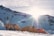 Bright sunlight illuminates snow-covered mountains with two orange tents pitched in the foreground. Several pairs of trekking poles and a helmet are positioned beside the tents, suggesting a camping or trekking expedition. The sky is partially cloudy, casting a dramatic light over the scene.