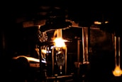 A dimly lit scene with a prominent glowing flame illuminating a workshop or industrial setting. Metallic structures and tubes are visible, suggesting a glassblowing process. The intense light creates sharp contrasts with the surrounding darkness.