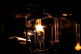 A dimly lit scene with a prominent glowing flame illuminating a workshop or industrial setting. Metallic structures and tubes are visible, suggesting a glassblowing process. The intense light creates sharp contrasts with the surrounding darkness.