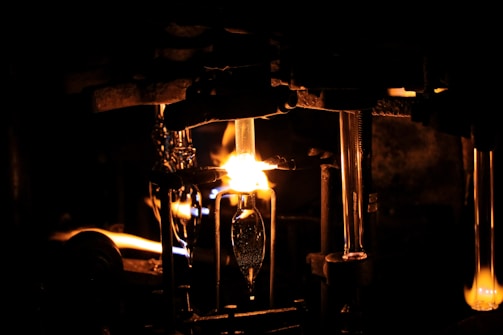 A dimly lit scene with a prominent glowing flame illuminating a workshop or industrial setting. Metallic structures and tubes are visible, suggesting a glassblowing process. The intense light creates sharp contrasts with the surrounding darkness.