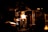 A dimly lit scene with a prominent glowing flame illuminating a workshop or industrial setting. Metallic structures and tubes are visible, suggesting a glassblowing process. The intense light creates sharp contrasts with the surrounding darkness.