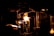 A dimly lit scene with a prominent glowing flame illuminating a workshop or industrial setting. Metallic structures and tubes are visible, suggesting a glassblowing process. The intense light creates sharp contrasts with the surrounding darkness.