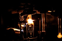 A dimly lit scene with a prominent glowing flame illuminating a workshop or industrial setting. Metallic structures and tubes are visible, suggesting a glassblowing process. The intense light creates sharp contrasts with the surrounding darkness.